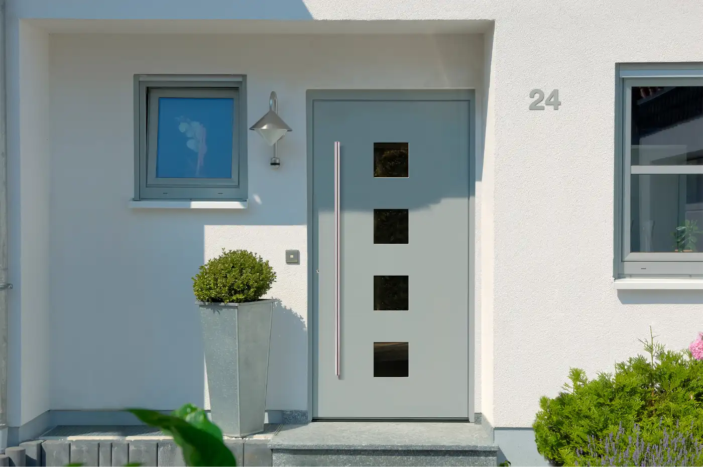 Hellgraue Haustür mit vier quadratischen Glasausschnitten und langem Edelstahlgriff an einer weißen Fassade